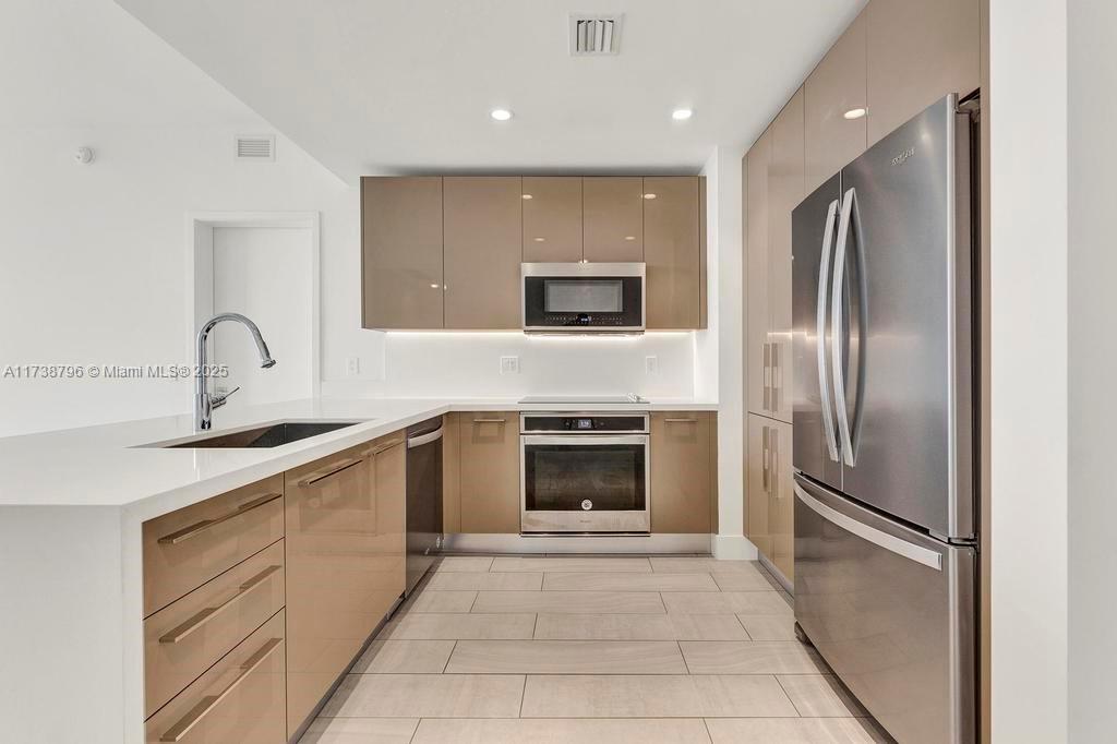 A modern kitchen features glossy beige cabinets, stainless steel appliances, and light tile flooring — 2000 Metropica Way # 2307, Sunrise FL 33323, SUNRISE, FL