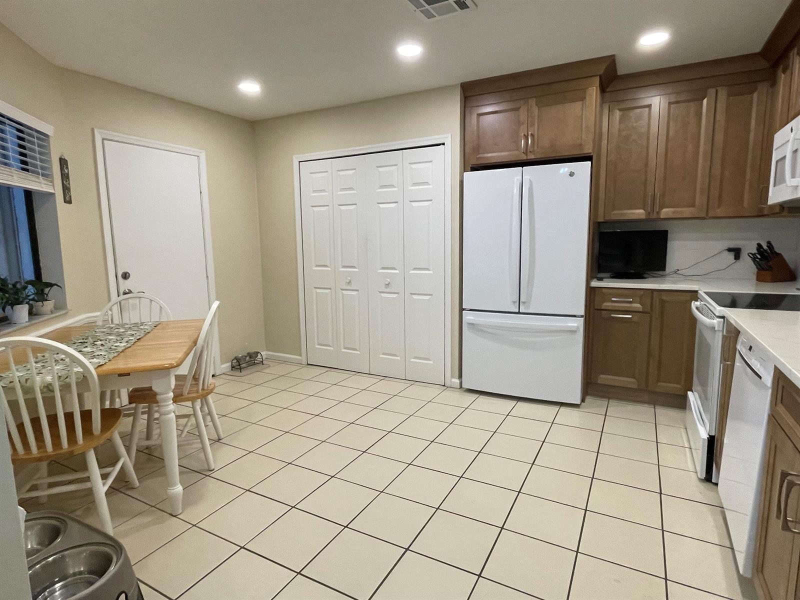 A kitchen with wood cabinets, white appliances, a dining table, and light-colored tile flooring — 124 Sandy Lane, Royal Palm Beach FL 33411, ROYAL PALM BEACH, FL