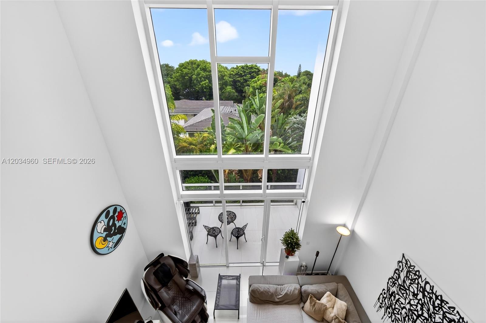 A high-ceilinged living room features floor-to-ceiling windows overlooking a balcony and lush greenery — 455 NE 39th # 311, Miami FL 33137, MIAMI, FL
