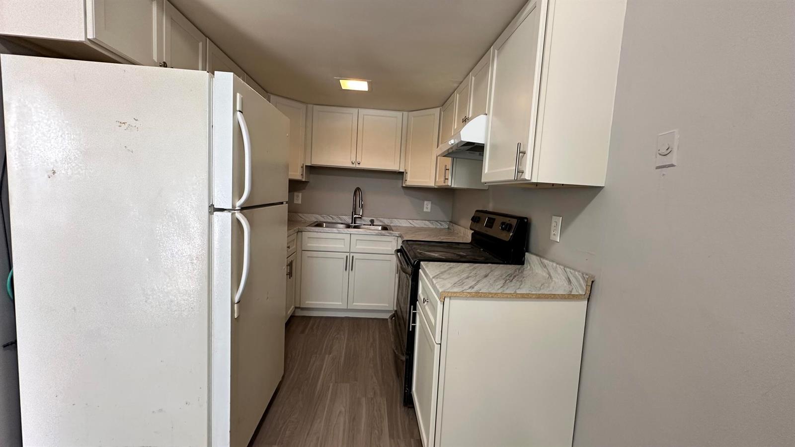 A galley kitchen features white cabinets, marble-look countertops, a white refrigerator, and wood-look flooring — 105 NE 6th Avenue, Boynton Beach FL 33435, BOYNTON BEACH, FL