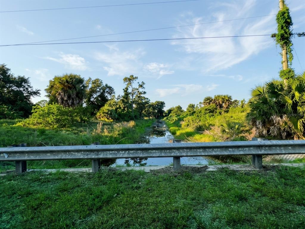 A metal guardrail overlooks a narrow canal surrounded by lush green vegetation and palm trees — 12323 CLARENDON AVENUE, PORT CHARLOTTE, FL
