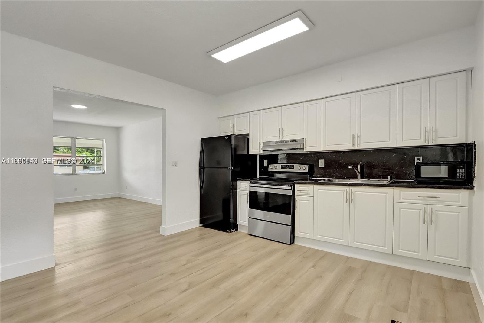 A kitchen with white cabinets, black countertops, and stainless steel appliances opens to a room — 1340 NW 102nd St, Miami FL 33147, MIAMI, FL