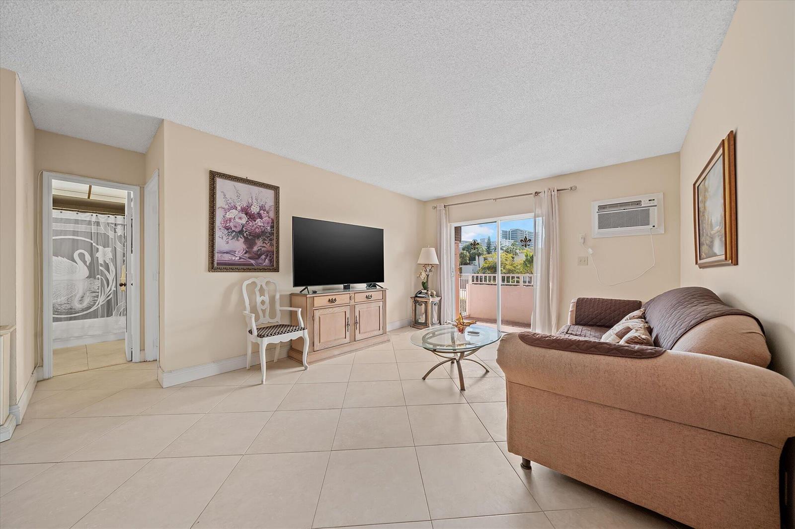 A living room with beige tile floors, a television on a console, and a sliding door — 3201 NE 14th Street Causeway # 209, Pompano Beach FL 33062, POMPANO BEACH, FL