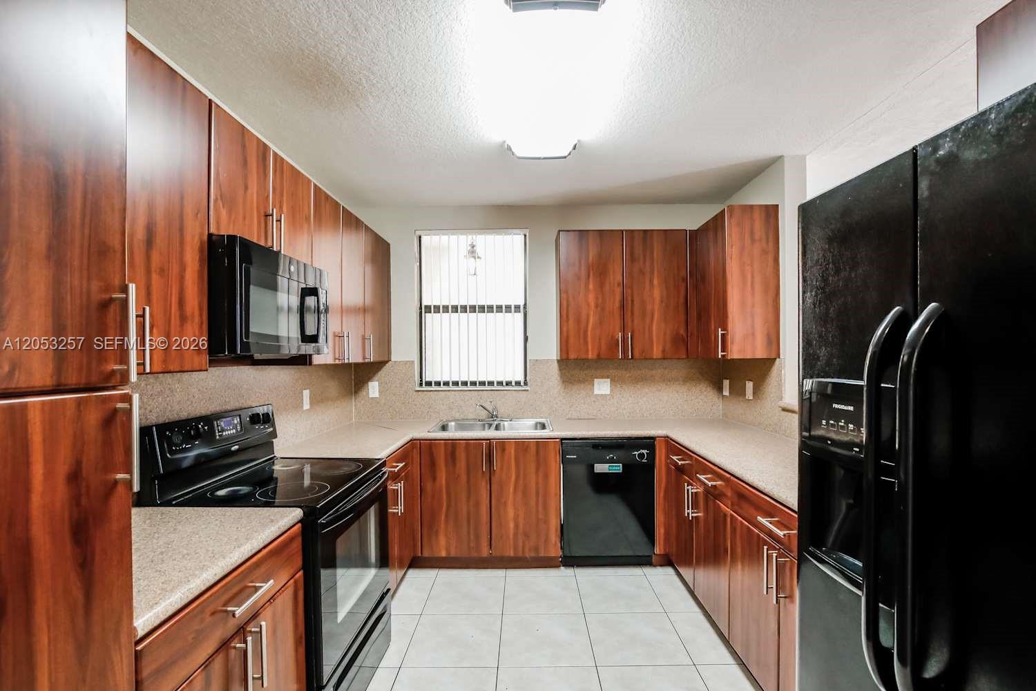 A kitchen features wood cabinets, black appliances, a double sink, and light-colored tile flooring — CUTLER BAY, FL