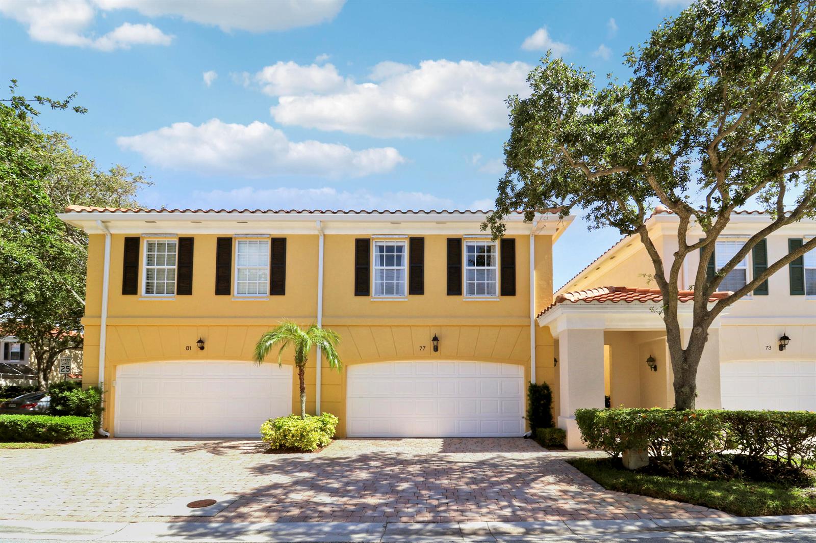 A yellow two-story townhouse exterior features two white garage doors and a paved driveway — 77 Laurel Oaks Circle, Tequesta FL 33469, TEQUESTA, FL