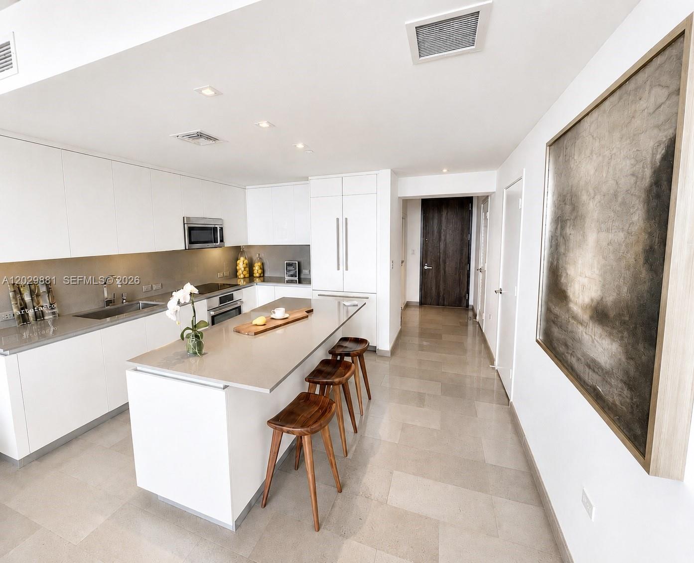 A modern white kitchen features a central island with wooden stools and stone countertops — 88 SW 7 # 3506, Miami FL 33130, MIAMI, FL