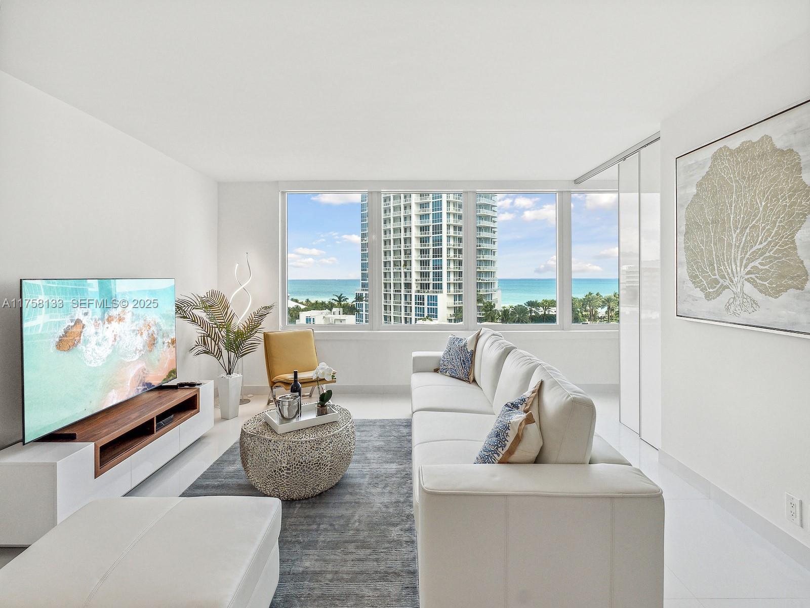 A bright living room features a white sofa, a television, and floor-to-ceiling windows overlooking the ocean — 400 S Pointe Dr # 906, Miami Beach FL 33139, MIAMI BEACH, FL