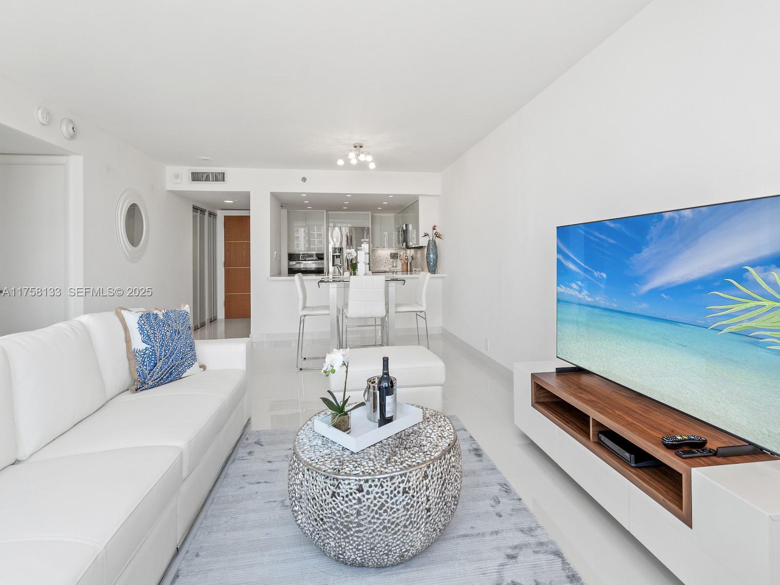 A bright living room features a white sofa, a textured coffee table, and a television — 400 S Pointe Dr # 906, Miami Beach FL 33139, MIAMI BEACH, FL