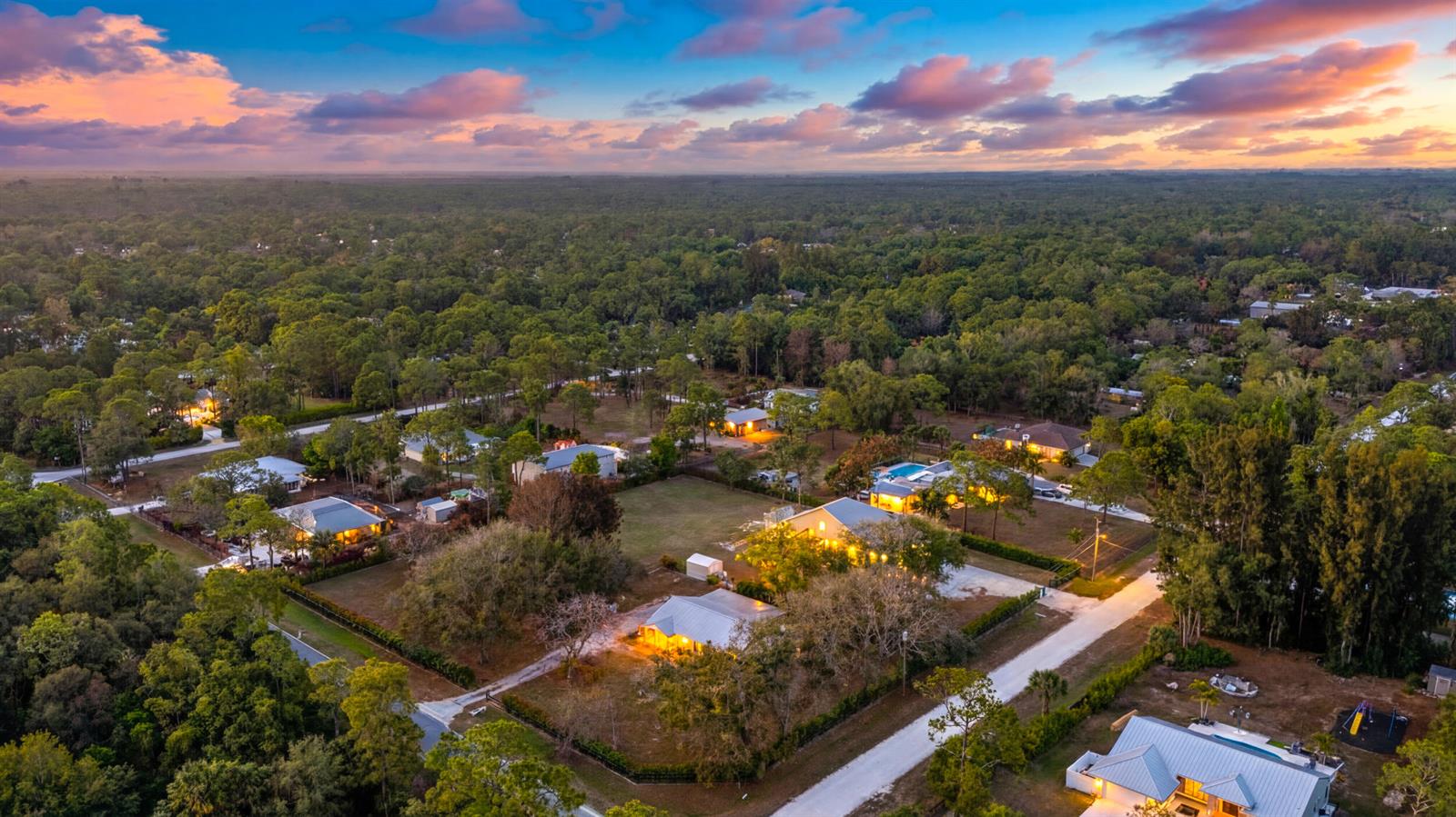 An aerial view shows several houses with illuminated windows nestled among trees at sunset — 15848 N 129th Pl N, FL 33478, FL