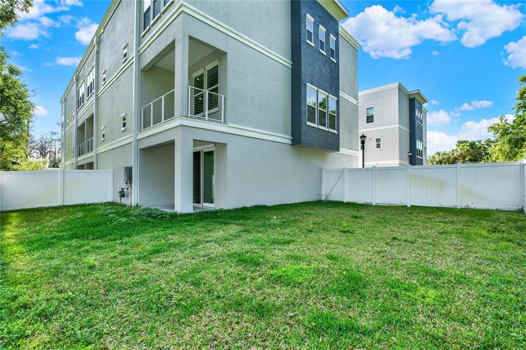 A view of a grassy backyard enclosed by a white fence behind a multi-story building — 3847 SEENA MANOR COURT, TAMPA, FL