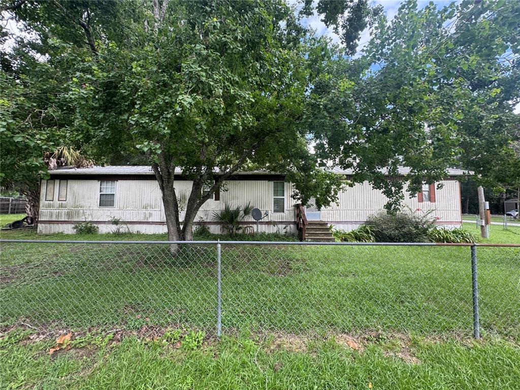 A single-story mobile home with white siding sits behind a chain-link fence and trees — 3285 SE 131ST PLACE, BELLEVIEW, FL
