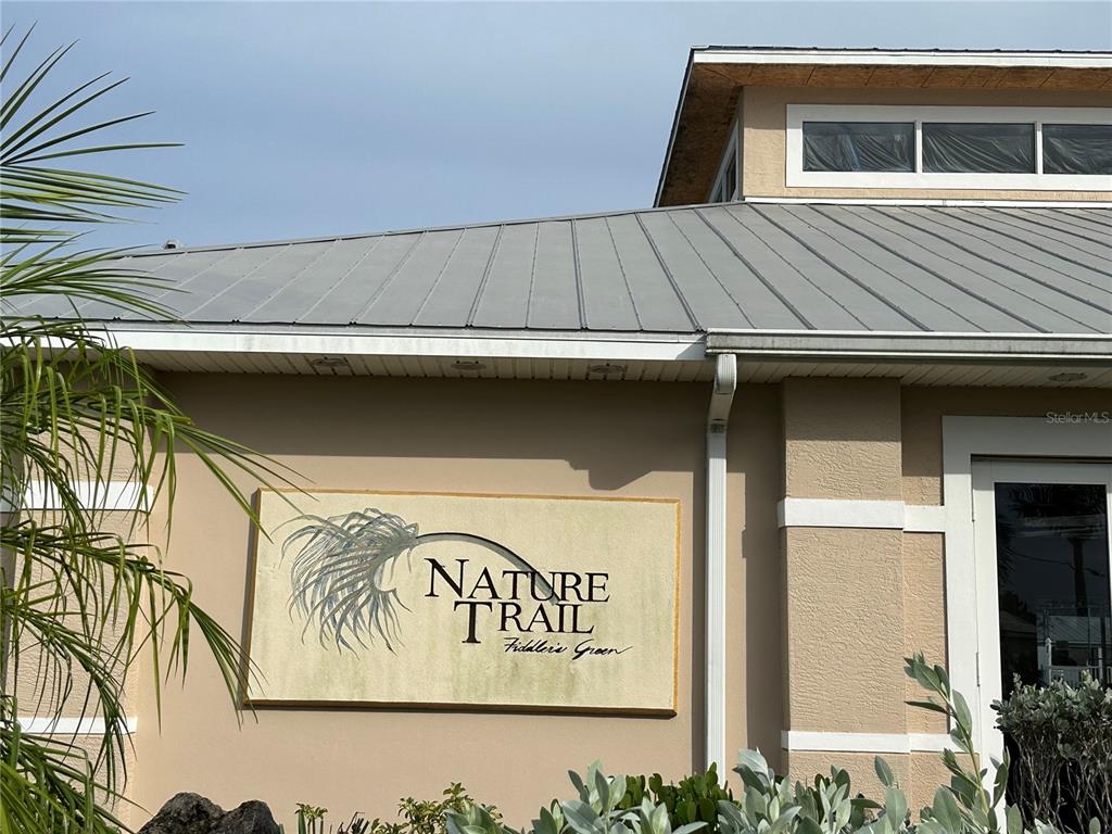 A tan building exterior features a metal roof and a sign reading Nature Trail Fiddler's Green — 9550 FIDDLERS GREEN CIRCLE UNIT 105, ROTONDA WEST, FL