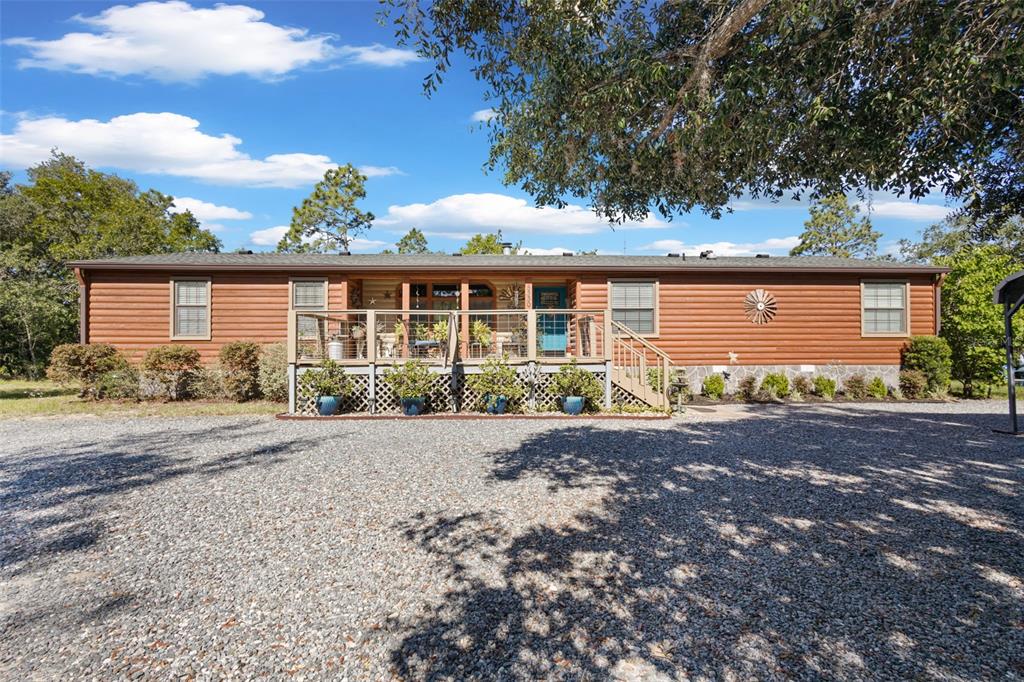 A single-story log-sided home features a front deck, gravel driveway, and surrounding trees — 5330 SE 128TH TERRACE, MORRISTON, FL
