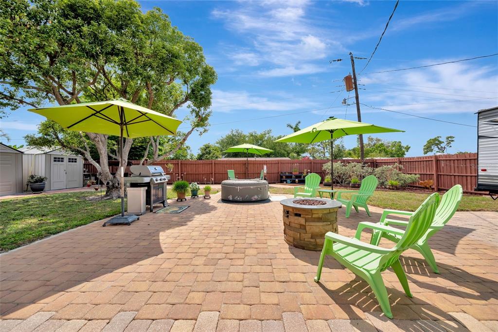 A backyard patio features a stone fire pit, green chairs, umbrellas, and a hot tub — 130 59TH STREET W, BRADENTON, FL