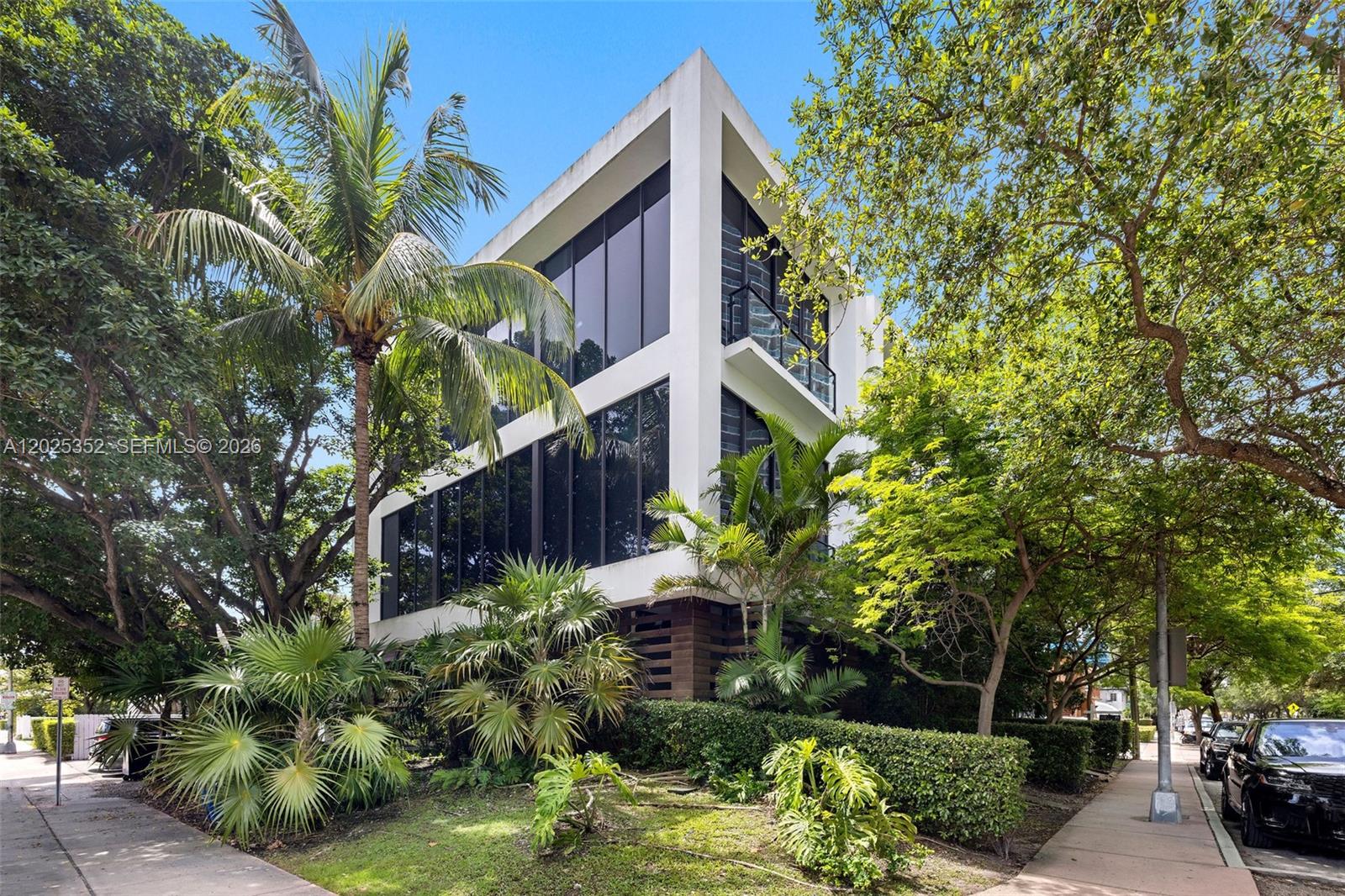 A modern three-story white building with floor-to-ceiling windows is surrounded by lush tropical landscaping — 1525 Bay Rd, Miami Beach FL 33139, MIAMI BEACH, FL