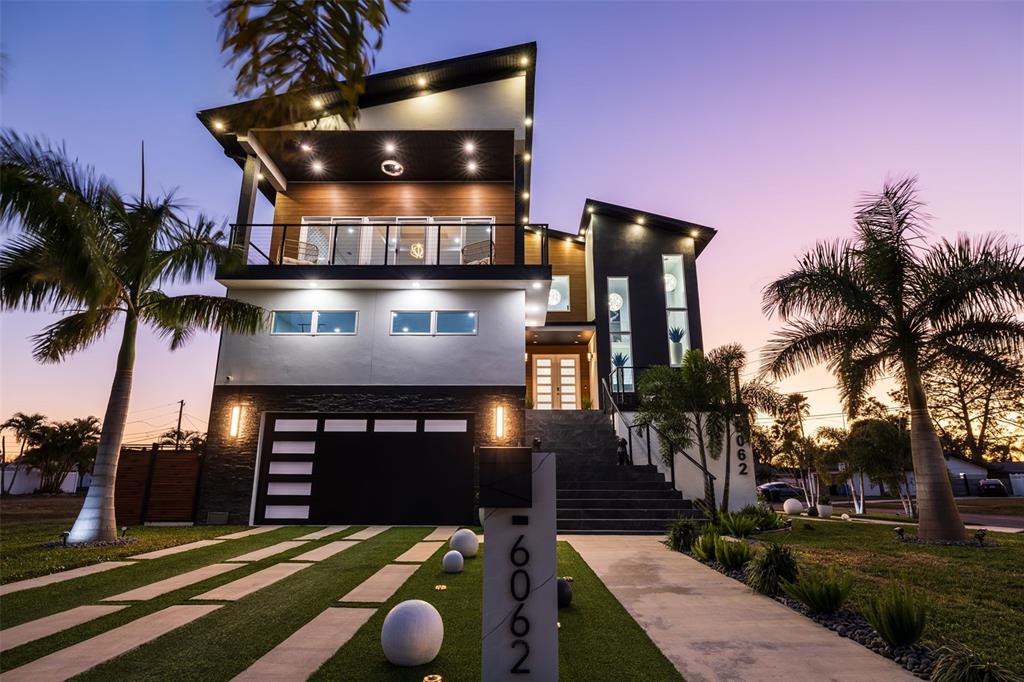 A modern three-story house with a garage, balcony, and palm trees at dusk — 6062 BAYOU GRANDE BOULEVARD NE, ST PETERSBURG, FL