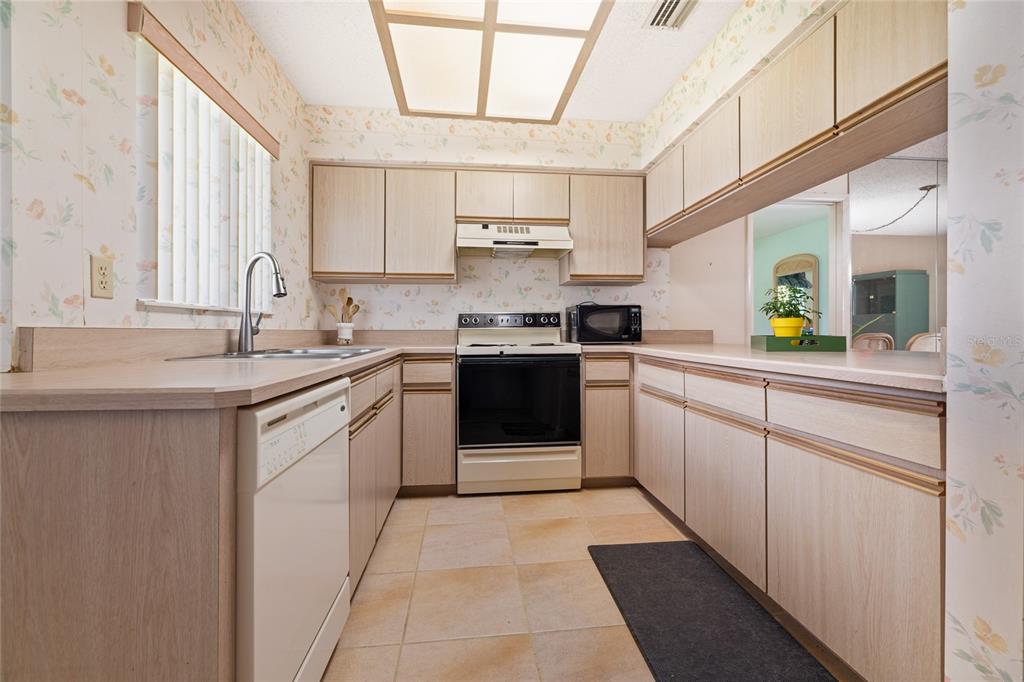 A kitchen features light wood cabinets, floral wallpaper, tile flooring, and a white dishwasher — 21 WEDGEWOOD LANE, PALM COAST, FL