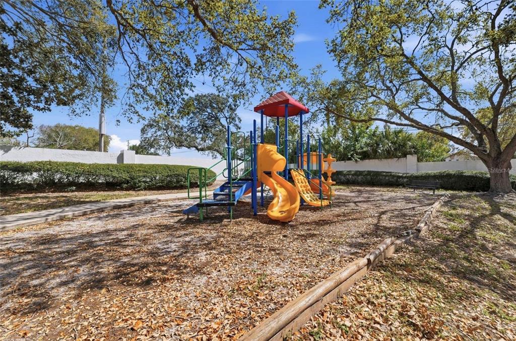A playground with a colorful slide structure sits on a wood-chipped lot under large trees — 3274 HAVILAND COURT UNIT 201, PALM HARBOR, FL