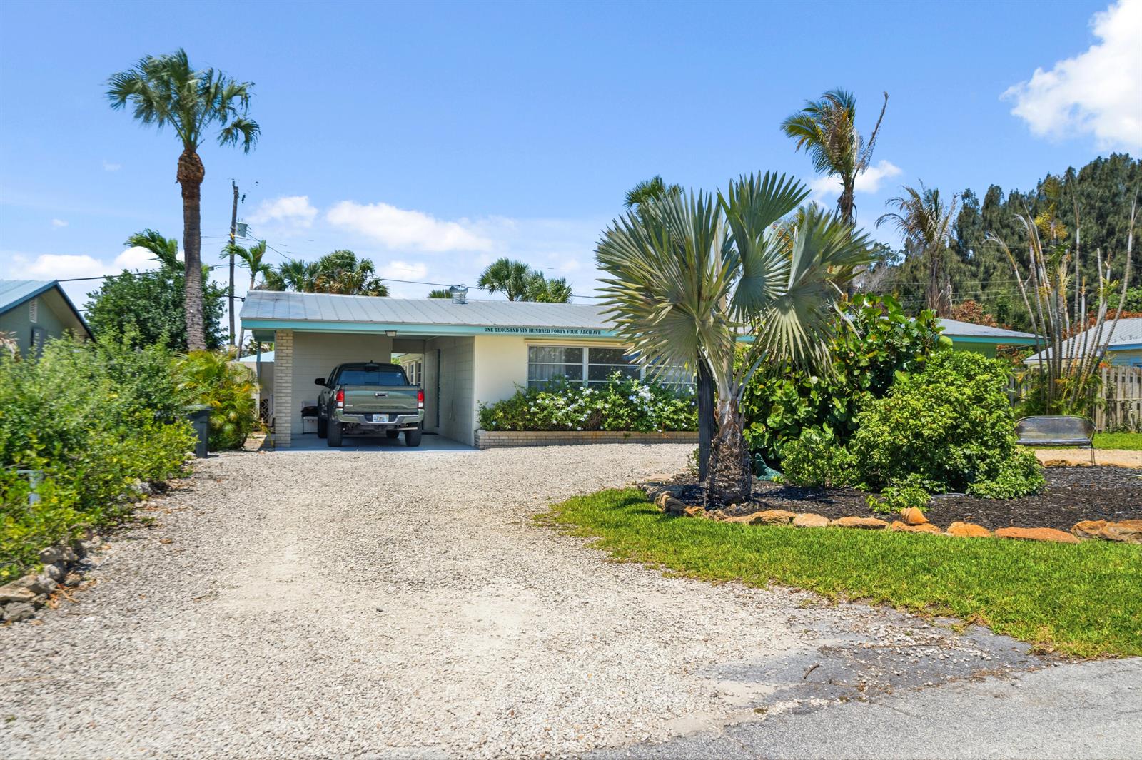 A single-story house with a gravel driveway, carport, and palm trees under a blue sky — 1644 NE Arch Avenue, Jensen Beach FL 34957, JENSEN BEACH, FL
