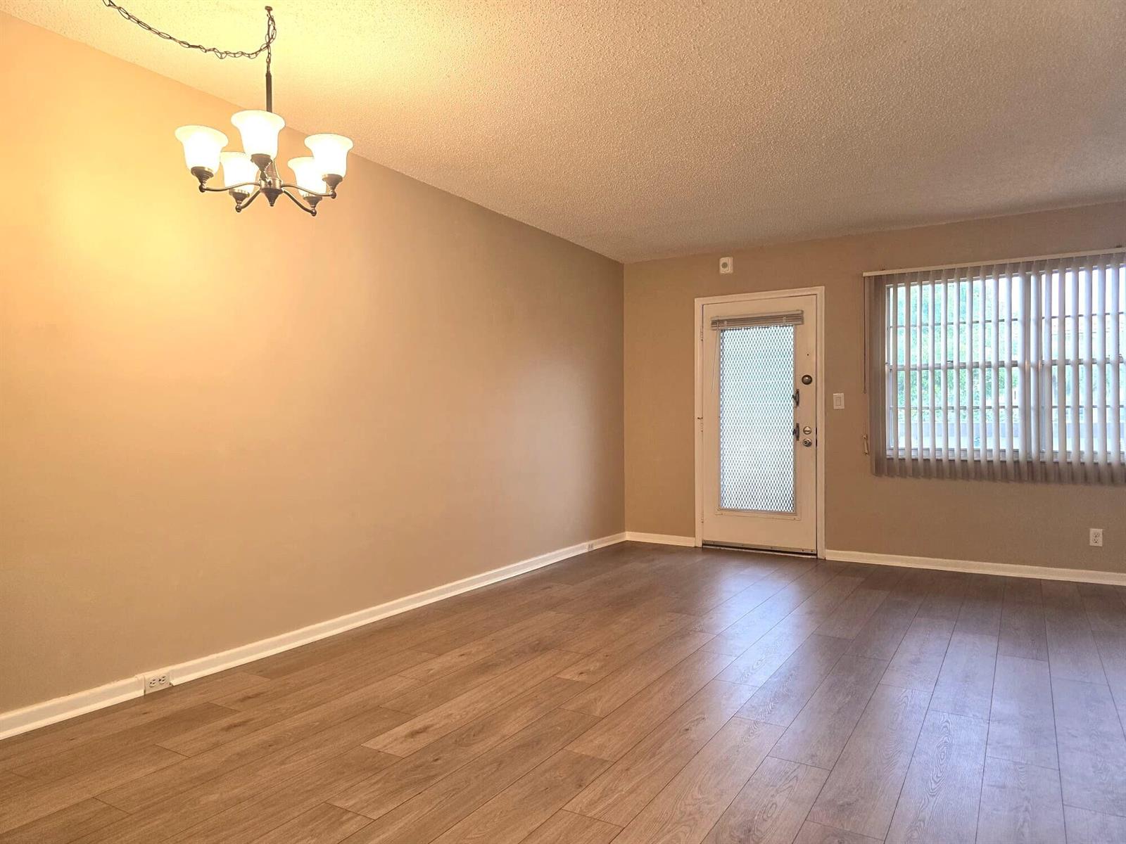 An empty living room features light brown wood-look flooring, beige walls, and a chandelier — 2103 Lucaya Bend # E2, Coconut Creek FL 33066, COCONUT CREEK, FL