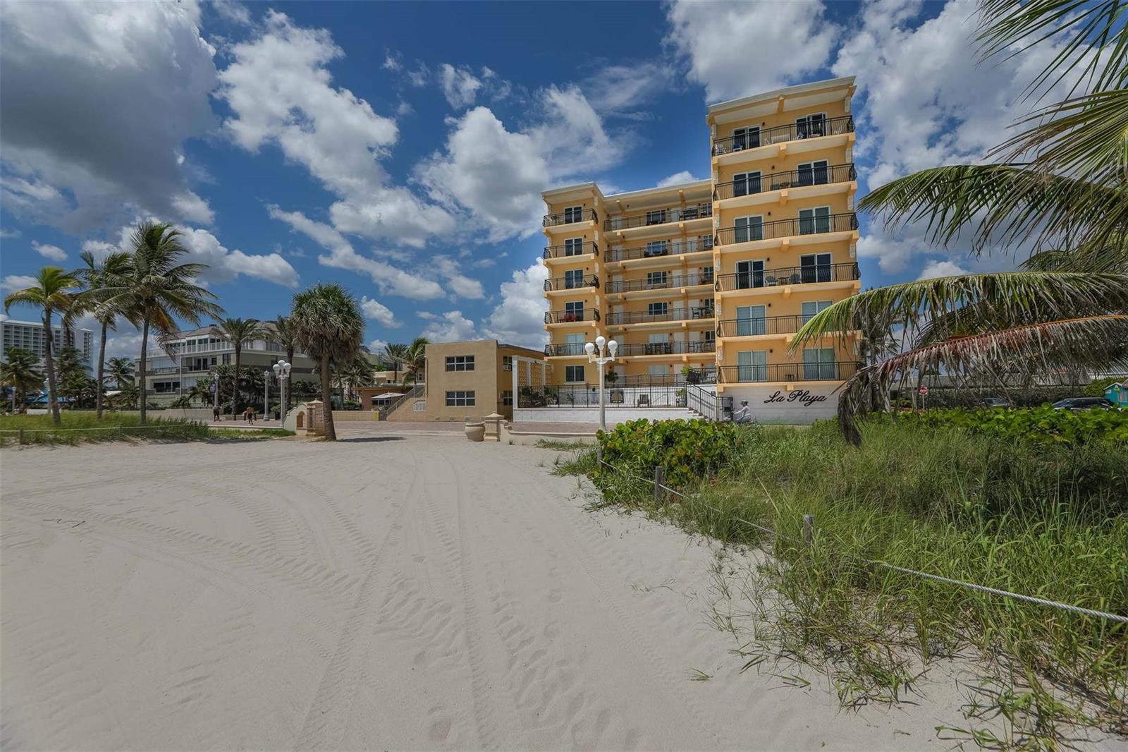 A multi-story yellow apartment building stands behind a sandy beach with palm trees — 1815 N Surf Road # 403, Hollywood FL 33019, HOLLYWOOD, FL