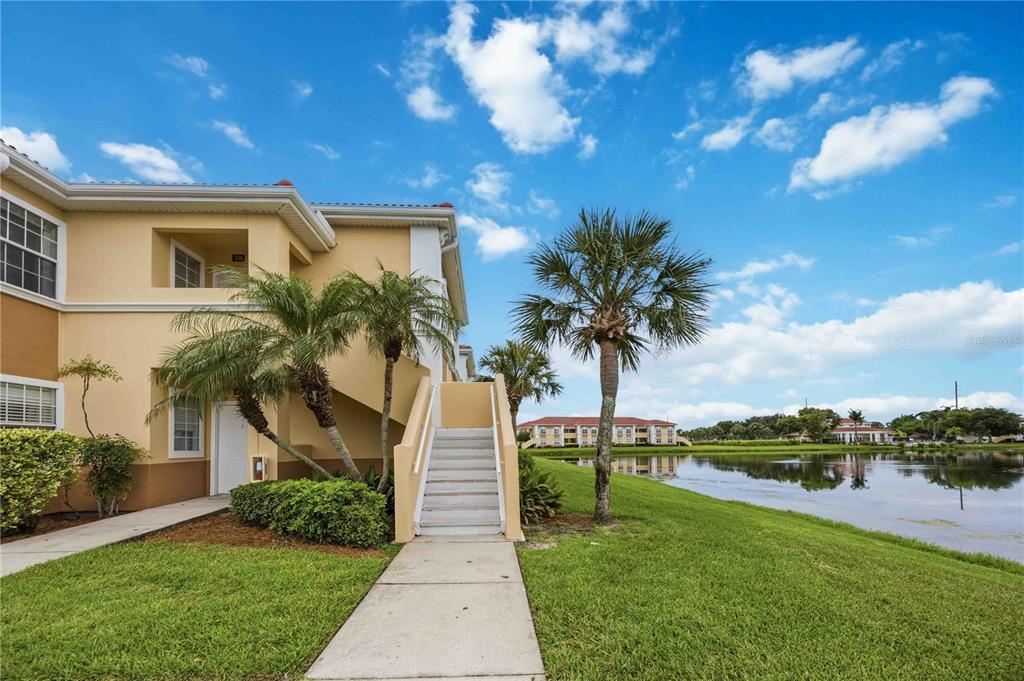 A two-story apartment building with exterior stairs sits beside a pond and palm trees — 1025 VILLAGIO CIRCLE UNIT 208, SARASOTA, FL