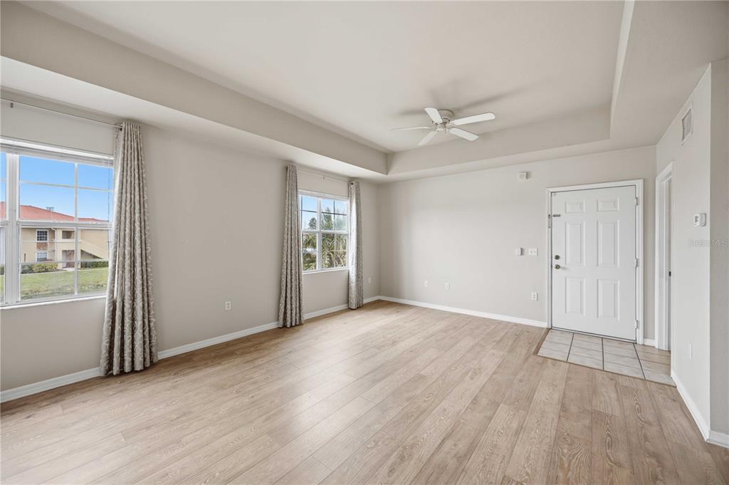 An empty living room with light wood-look flooring, a tray ceiling, and two windows — 1025 VILLAGIO CIRCLE UNIT 208, SARASOTA, FL