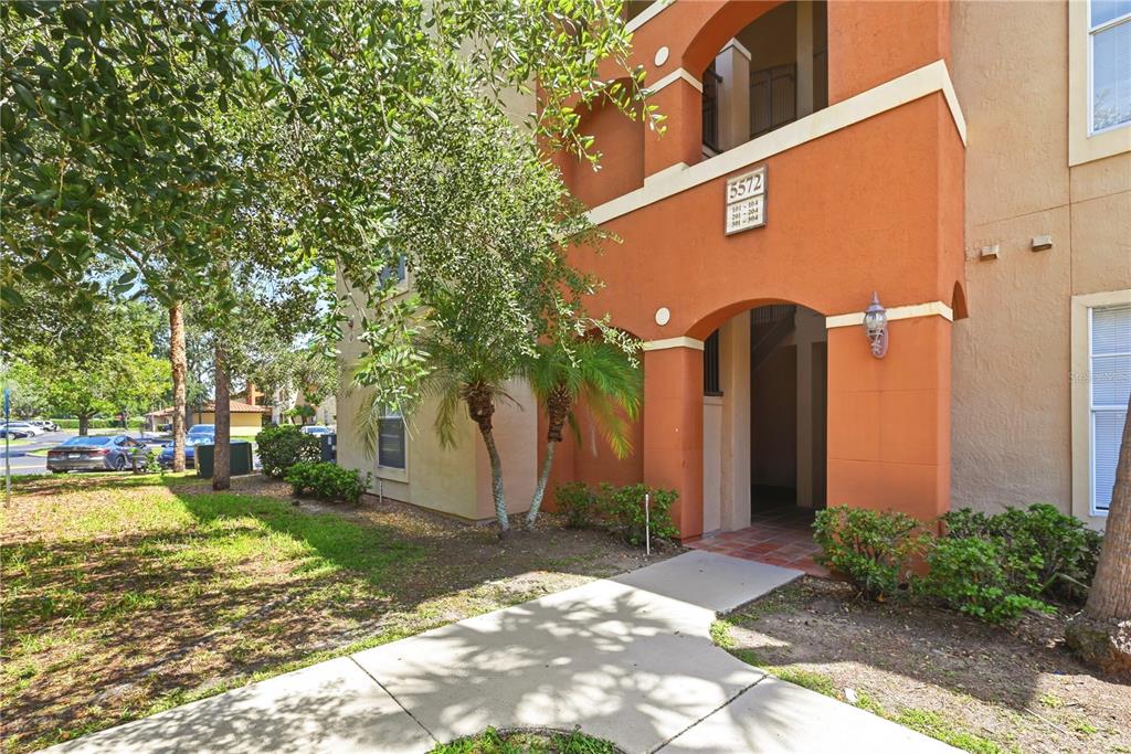 A sidewalk leads to the arched entrance of a multi-story orange stucco apartment building — 5572 METROWEST BOULEVARD UNIT 5-304, ORLANDO, FL