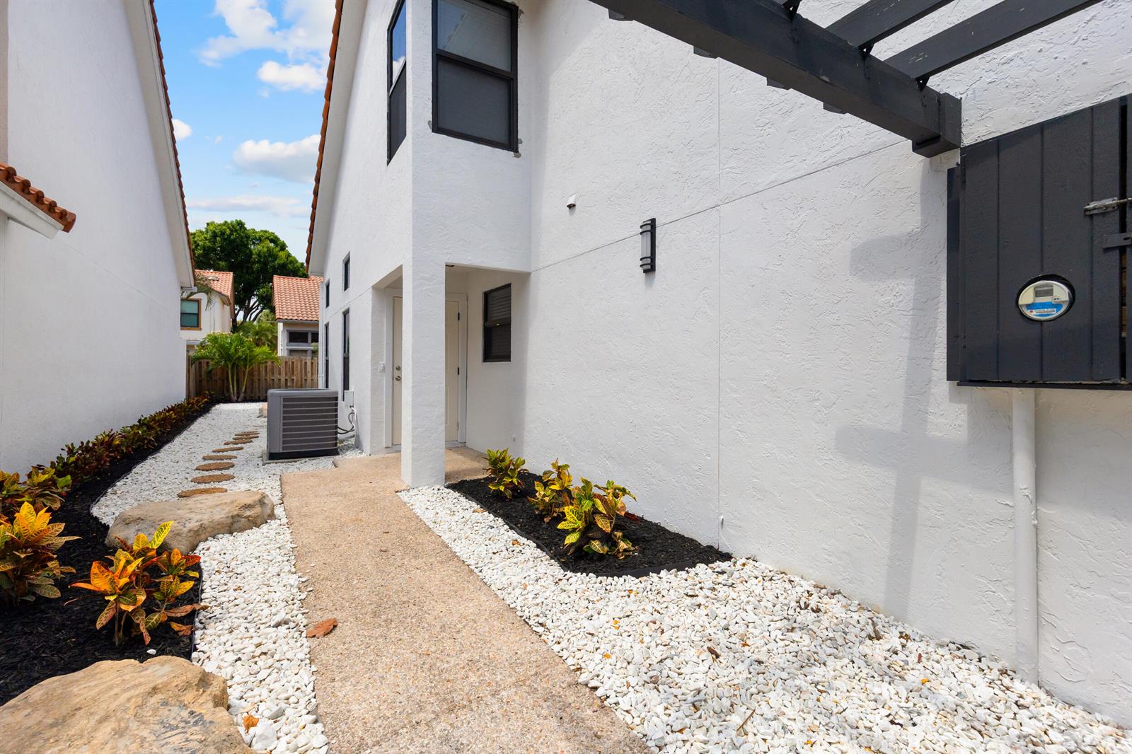 A white stucco exterior wall features a concrete walkway, landscaping, and an outdoor utility meter — 5521 Ilford Court, Boca Raton FL 33486, BOCA RATON, FL