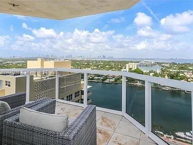 A high-rise balcony with wicker furniture overlooks a waterway and a distant city skyline — 4775 Collins Ave # 2105, Miami Beach FL 33140, MIAMI BEACH, FL