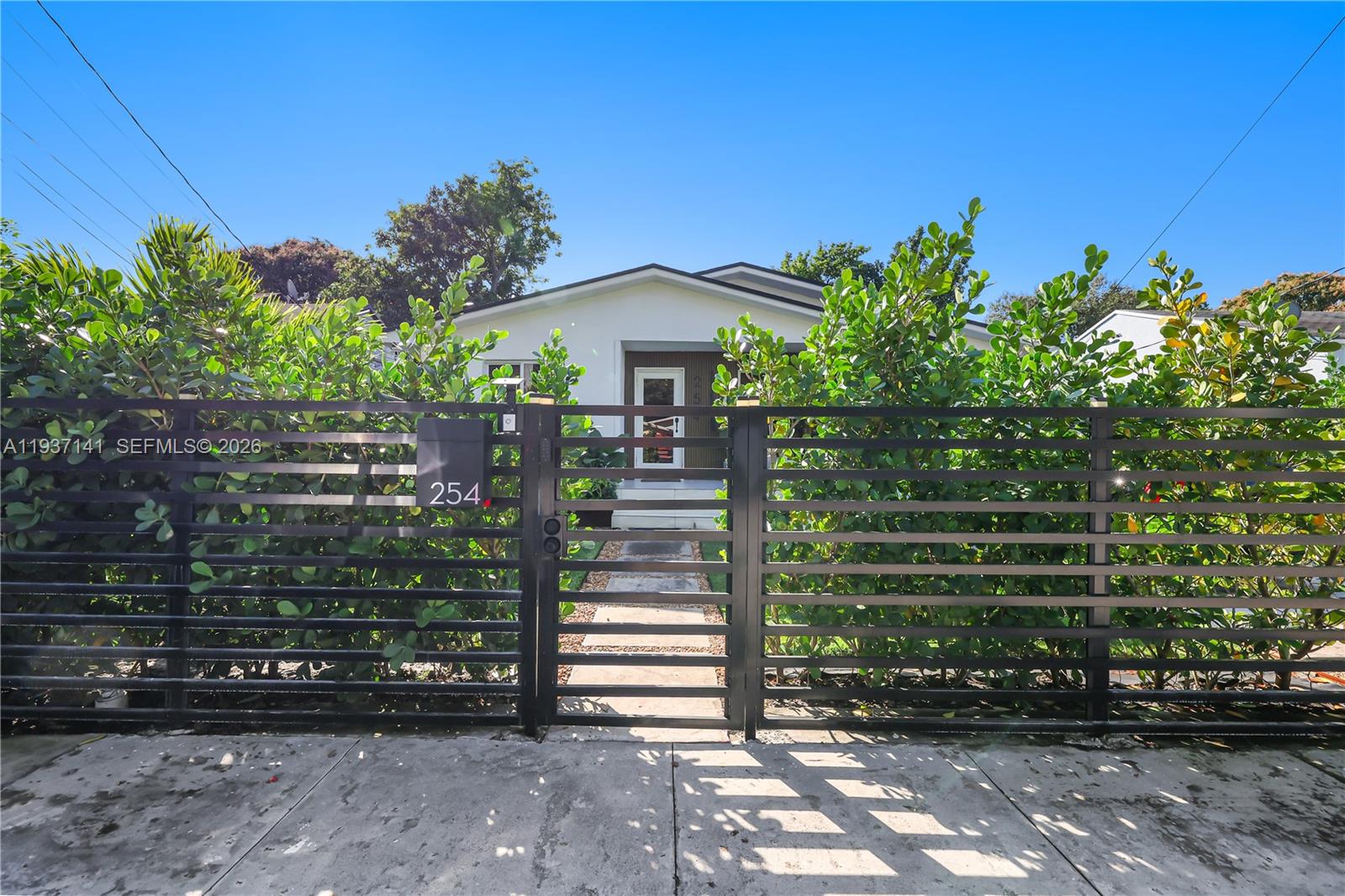 A white house is partially obscured by lush green hedges behind a black metal fence — 254 NW 43rd St, Miami FL 33127, MIAMI, FL