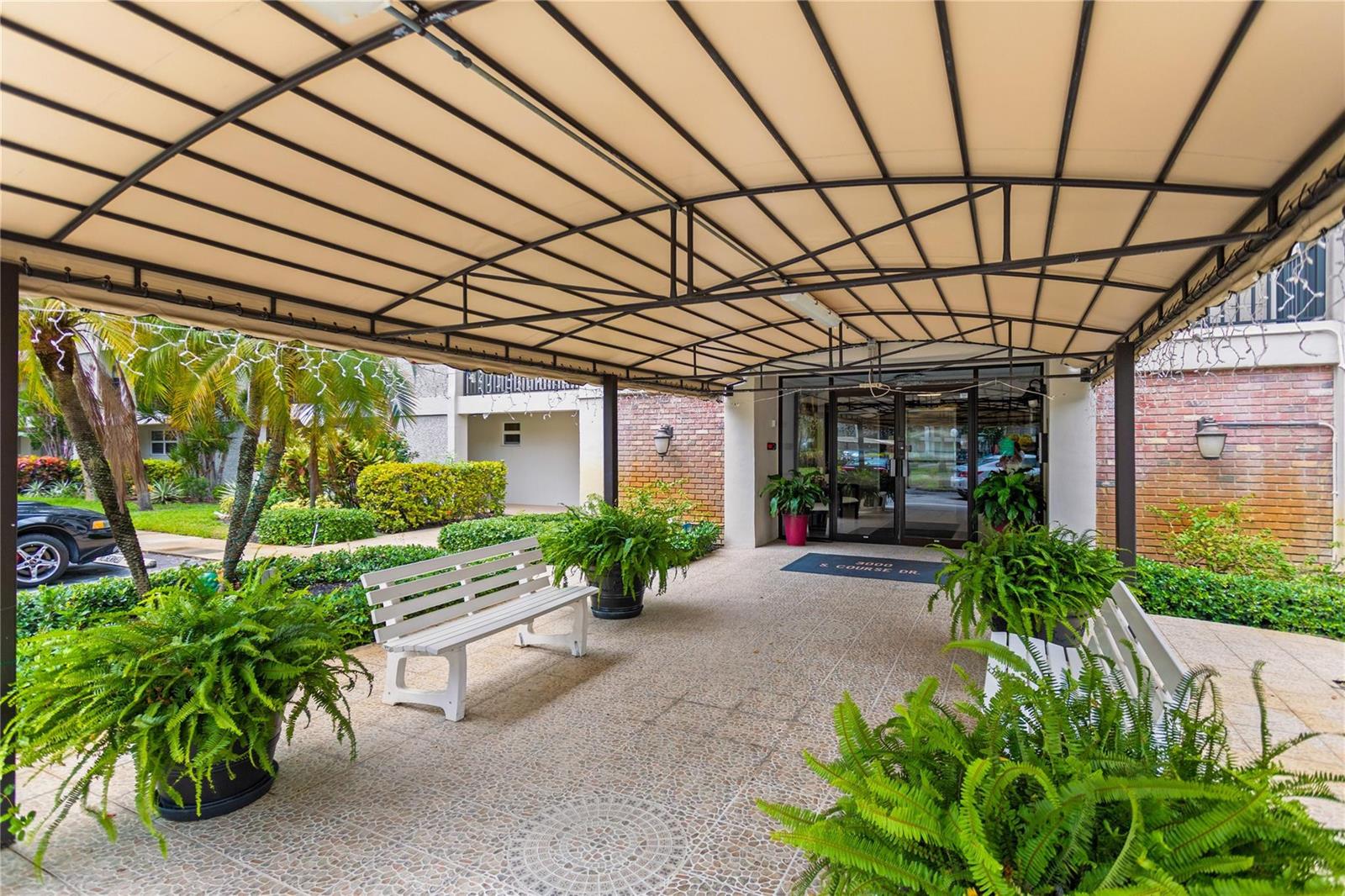 A covered entrance features a paved walkway, two white benches, and several potted ferns — 3000 S Course Drive # 607, Pompano Beach FL 33069, POMPANO BEACH, FL