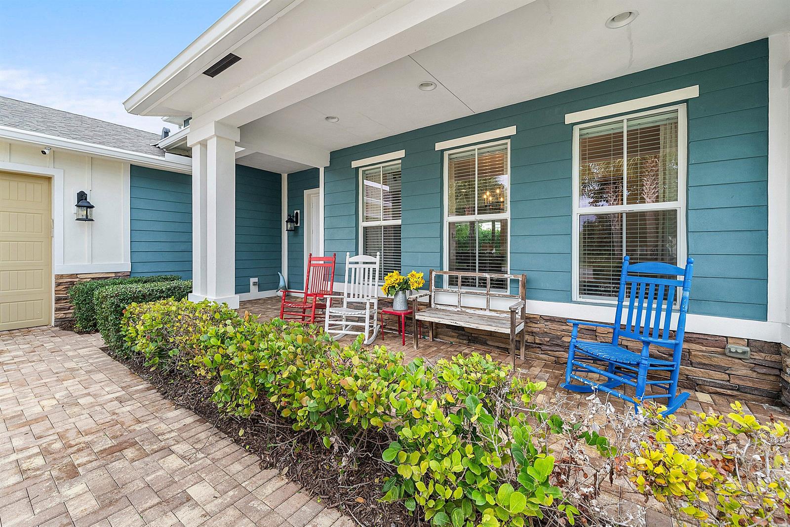 A covered porch features blue siding, stone accents, brick pavers, and several colorful rocking chairs — 1107 Timber Reap Trail, Loxahatchee FL 33470, LOXAHATCHEE, FL