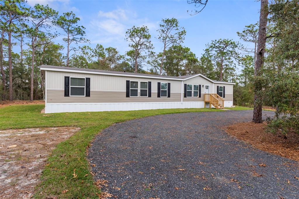 A single-story manufactured home with beige siding sits behind a curved gravel driveway — 6149 LA PINE ROAD, BROOKSVILLE, FL