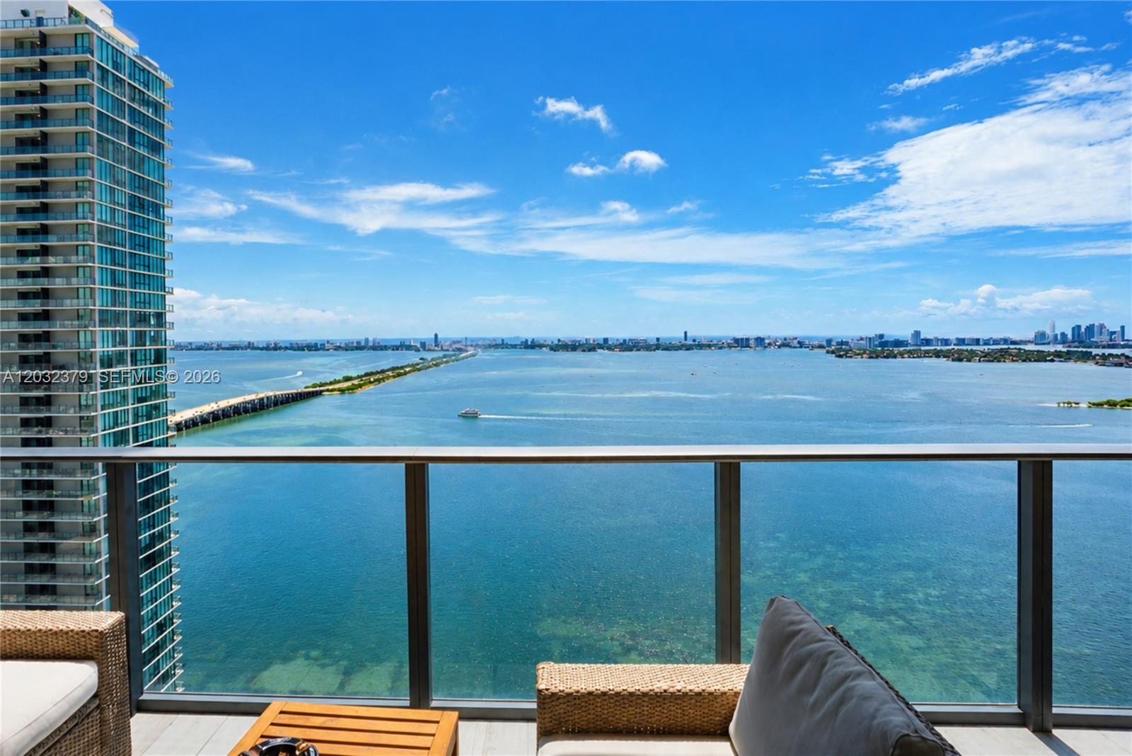 A balcony with wicker furniture overlooks a large bay and a distant city skyline — 480 NE 31st St # 3705, Miami FL 33137, MIAMI, FL