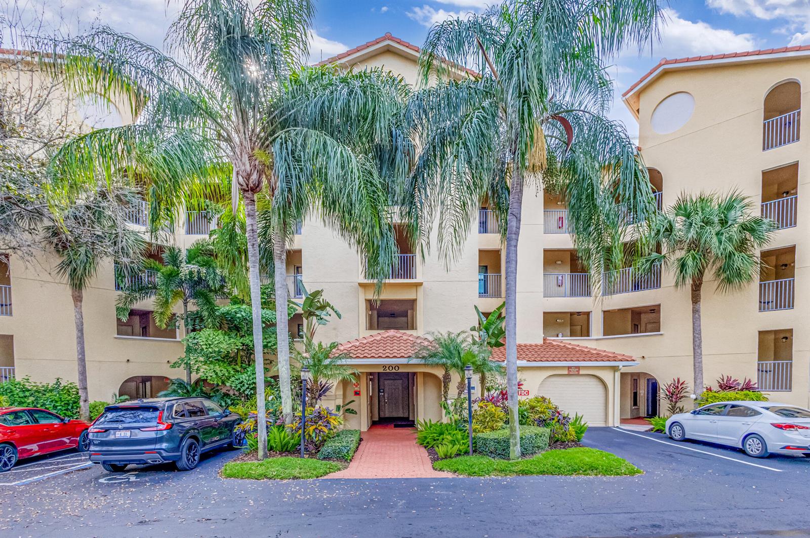 The exterior of a multi-story apartment building features palm trees, a tiled entrance, and parking — 200 Uno Lago Drive # 105, Juno Beach FL 33408, JUNO BEACH, FL