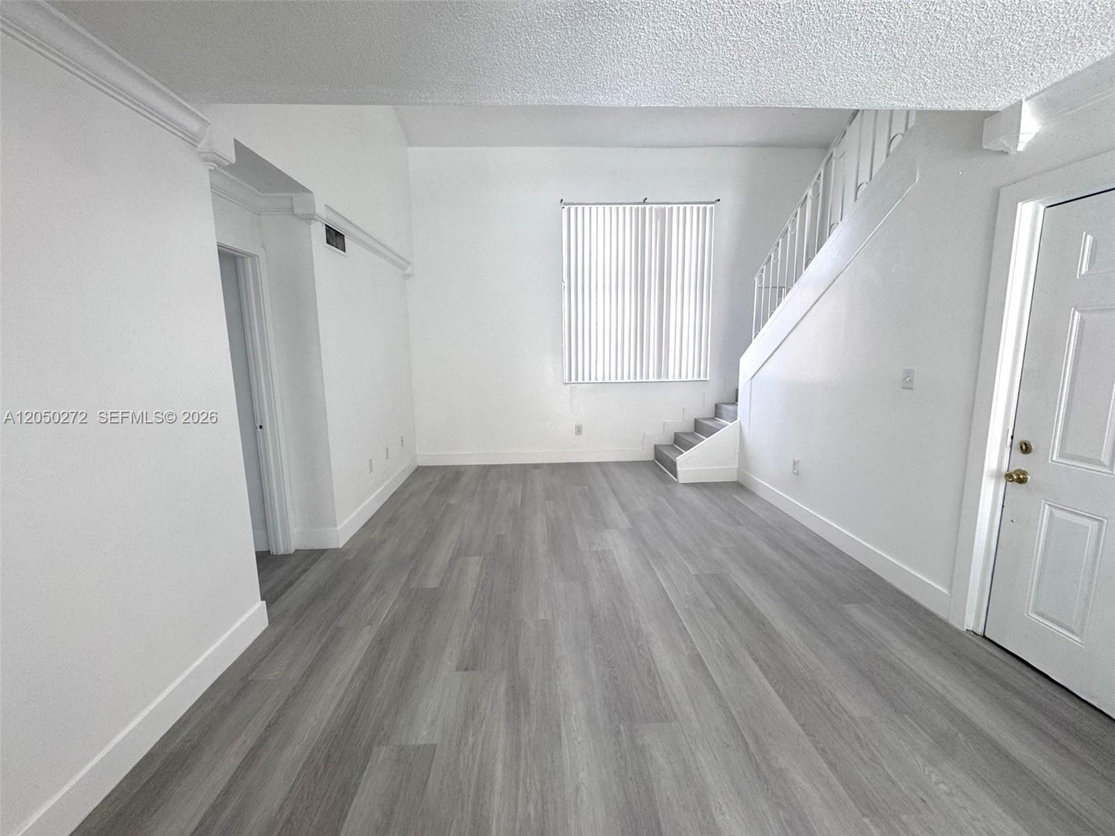 An empty room features white walls, gray wood-look flooring, a staircase, and vertical blinds — 2740 W 62nd St # 205, Hialeah FL 33016, HIALEAH, FL