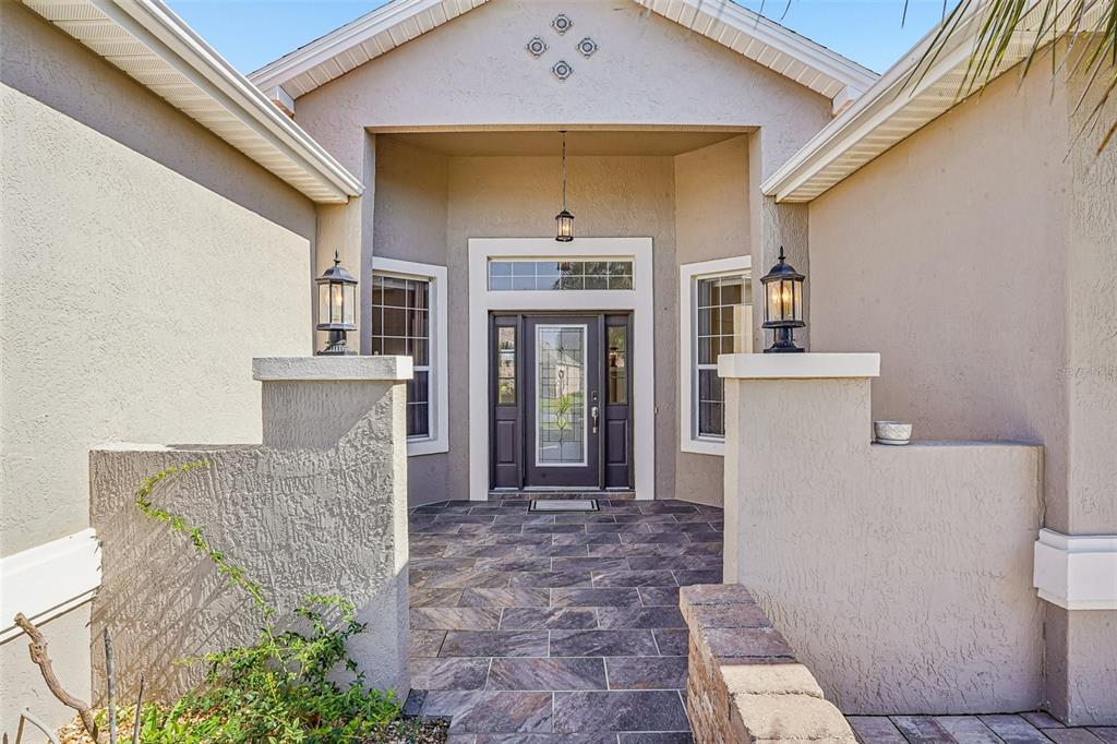 A covered entryway features a dark-framed glass door, stone-look tile flooring, and two pillar-mounted lanterns — 2013 SANTO DOMINGO DRIVE, THE VILLAGES, FL