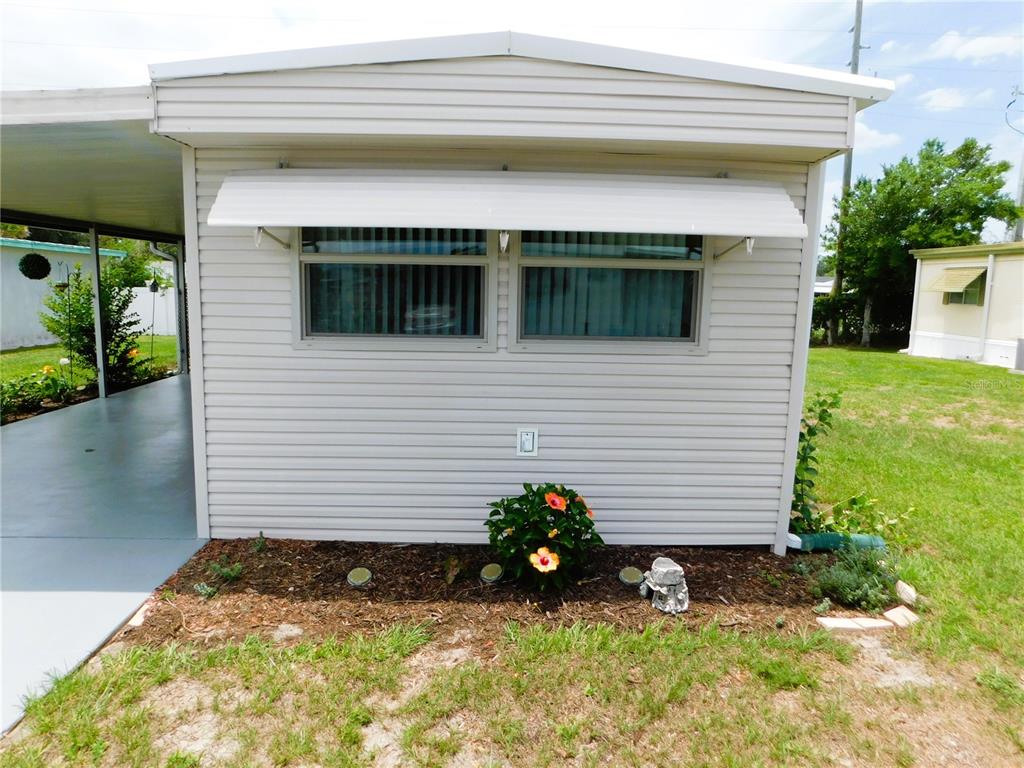 The exterior of a light-colored mobile home features horizontal siding, a window awning, and landscaping — 5315 ANTIGUA DRIVE, ZEPHYRHILLS, FL