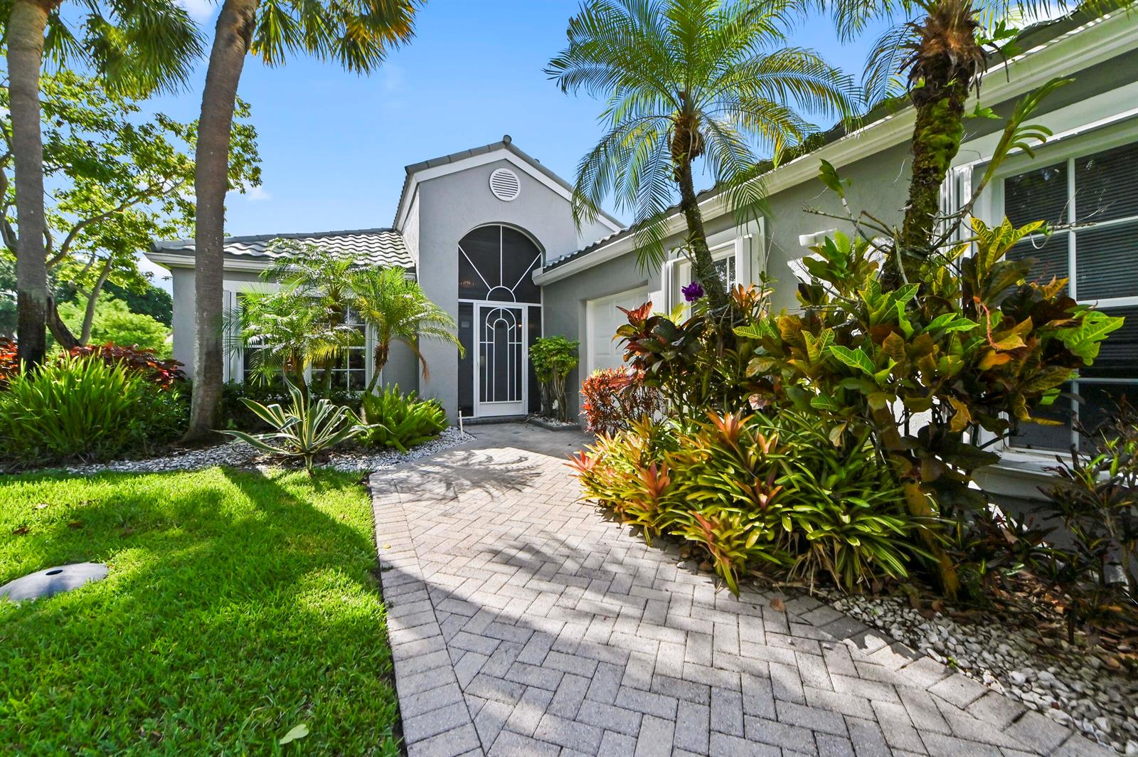 A paved walkway leads to the front entrance of a gray house with tropical landscaping — 6226 Water Lilly Lane, Boynton Beach FL 33437, BOYNTON BEACH, FL