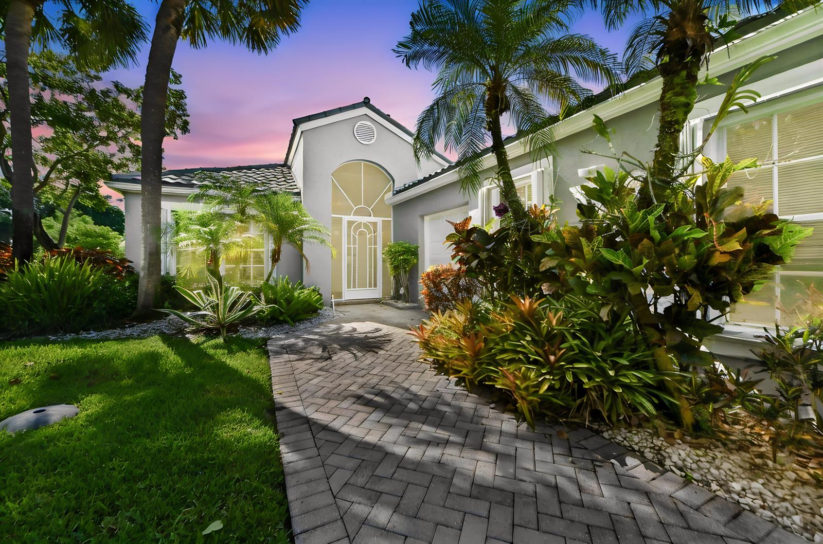 A paved walkway leads to the arched entryway of a gray house with lush landscaping — 6226 Water Lilly Lane, Boynton Beach FL 33437, BOYNTON BEACH, FL