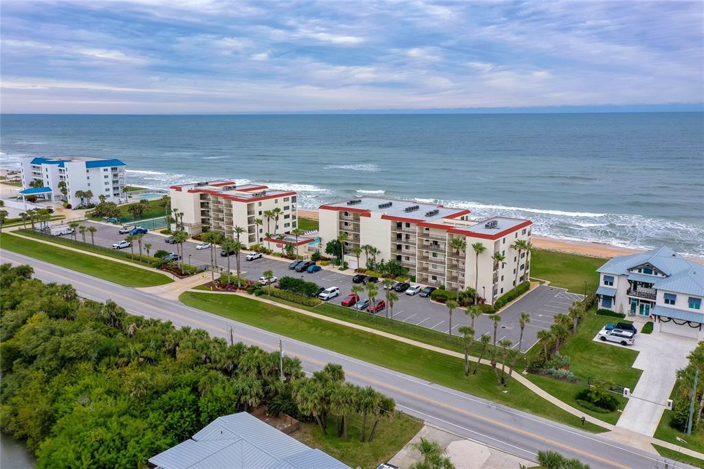 An aerial view shows beachfront apartment buildings with parking lots and a nearby coastal road — 6727 TURTLEMOUND ROAD UNIT 315, NEW SMYRNA BEACH, FL