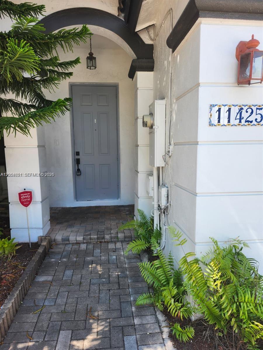 A gray front door is set within a covered entryway featuring brick pavers and ferns — 11425 NW 76th Ter, Doral FL 33178, DORAL, FL