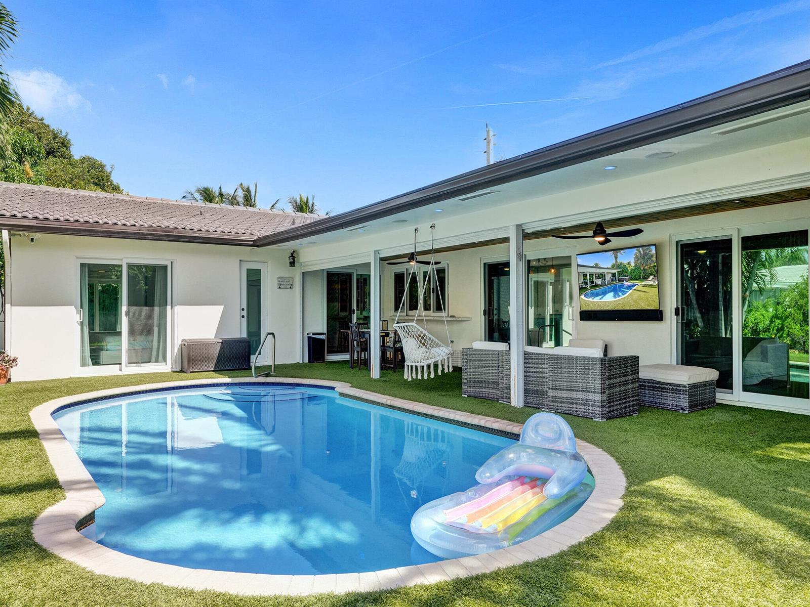 A backyard patio features a swimming pool, artificial turf, outdoor seating, and a wall-mounted television — 3821 NE 29th Avenue, Lighthouse Point FL 33064, LIGHTHOUSE POINT, FL
