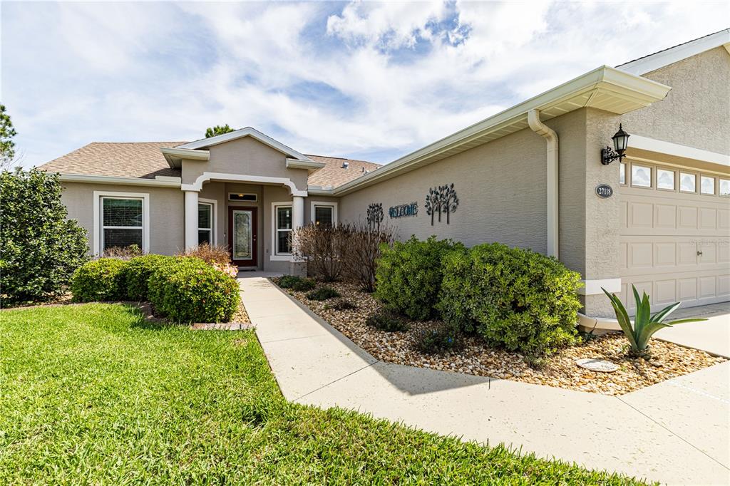 A single-story house exterior features a beige stucco facade, a concrete walkway, and a garage — 27118 ROANOKE DRIVE, LEESBURG, FL