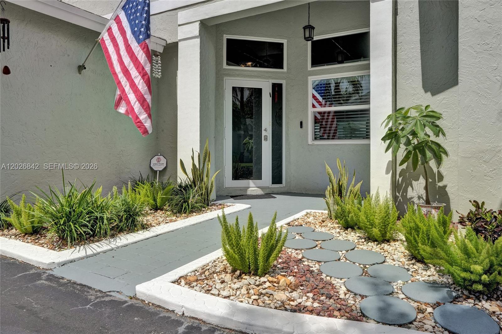 The exterior entrance features a walkway, landscaping, a flag, and a glass-paneled front door — 10639 Lago Welleby Dr, Sunrise FL 33351, SUNRISE, FL