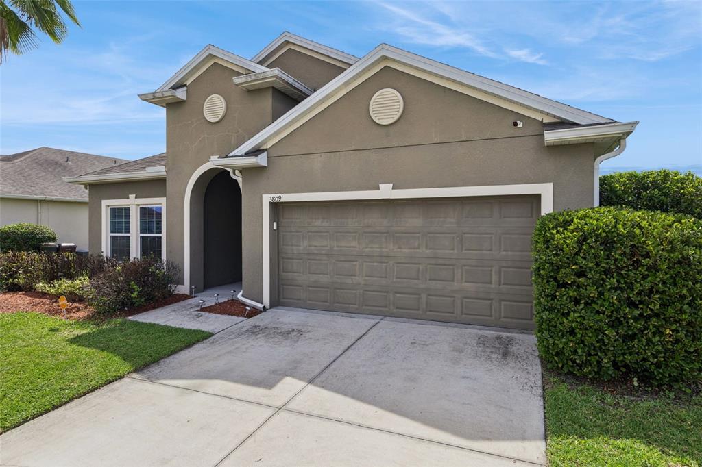 A tan stucco house features a two-car garage, a covered entryway, and a concrete driveway — 3809 ENCHANTMENT LANE, ST CLOUD, FL