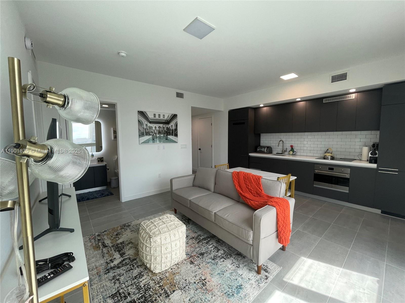 A modern studio apartment features a grey sofa, kitchen cabinetry, and tile flooring — 227 NE 2nd St # 1606, Miami FL 33132, MIAMI, FL