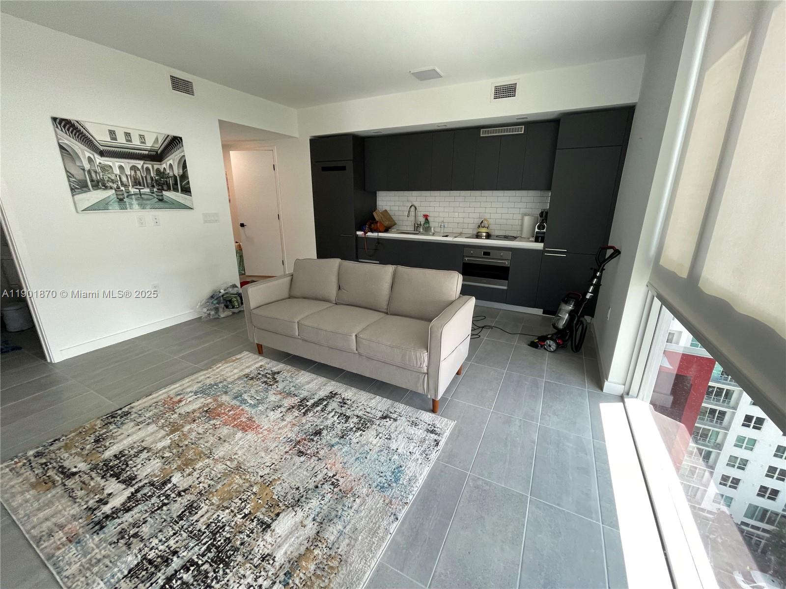 A studio apartment features a gray sofa, a patterned rug, and a dark kitchen — 227 NE 2nd St # 1606, Miami FL 33132, MIAMI, FL