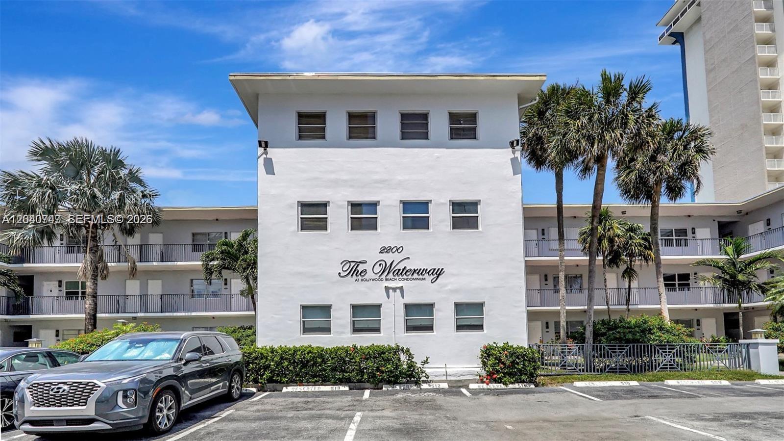 A three-story white apartment building features a parking lot and palm trees in front — HOLLYWOOD, FL